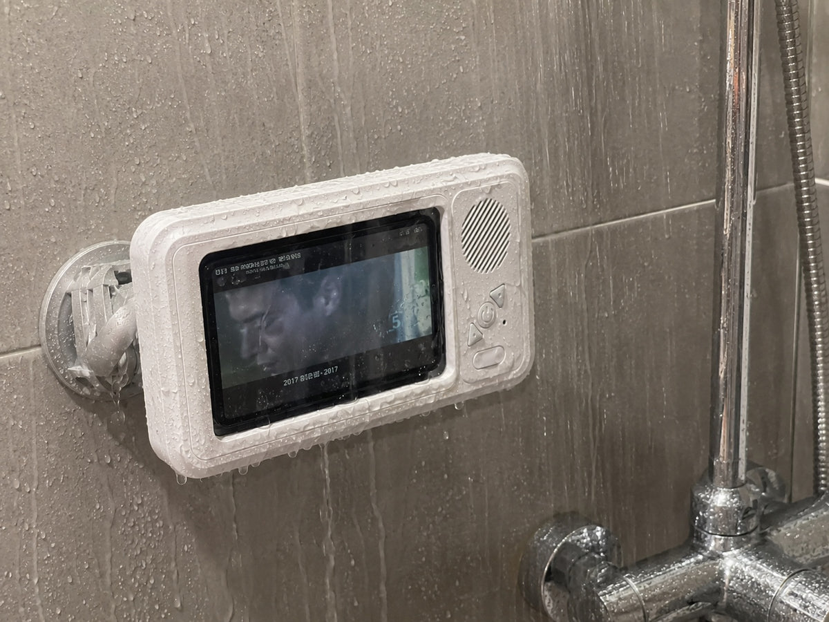 A white waterproof phone holder displaying a dark-haired man on screen is mounted with a suction cup on a wet grey tiled shower wall next to a chrome shower head and mixer, all covered in water droplets